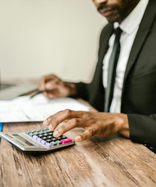 A businessman in a black suit using a calculator at a desk with financial documents and a laptop.