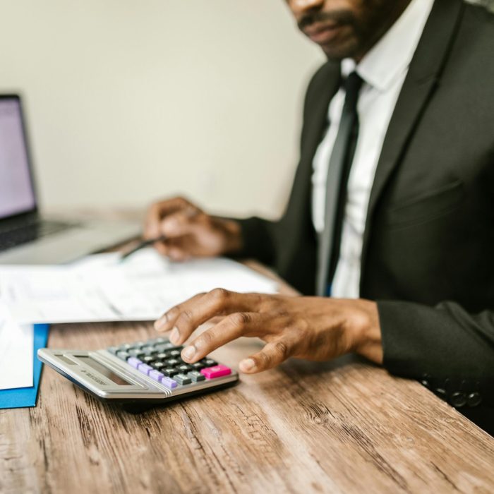 A businessman in a black suit using a calculator at a desk with financial documents and a laptop.